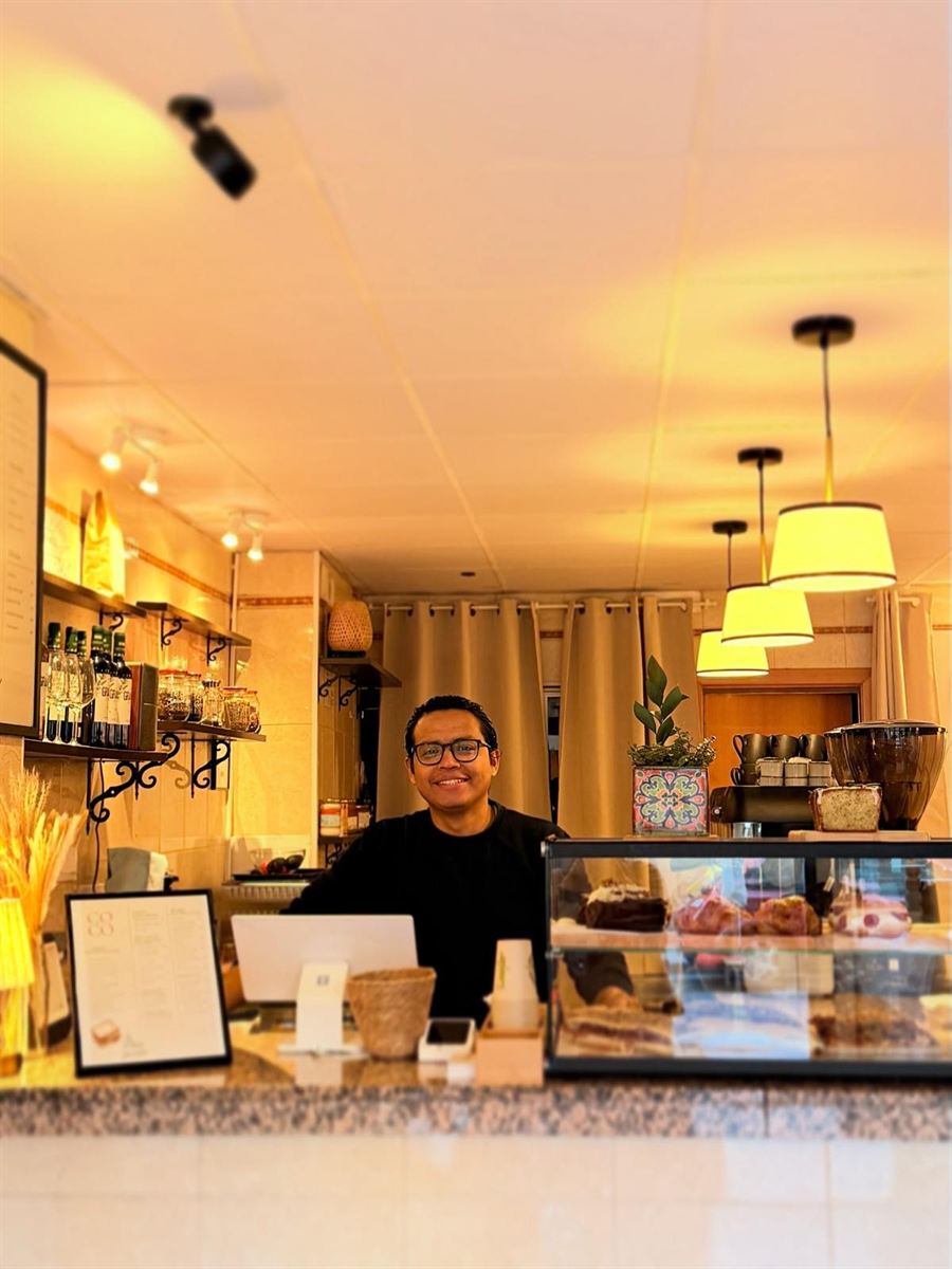 Uno de los fundadores de Lúcido sonriendo detrás de la barra, con las lámparas encendidas y la vitrina de repostería al lado.