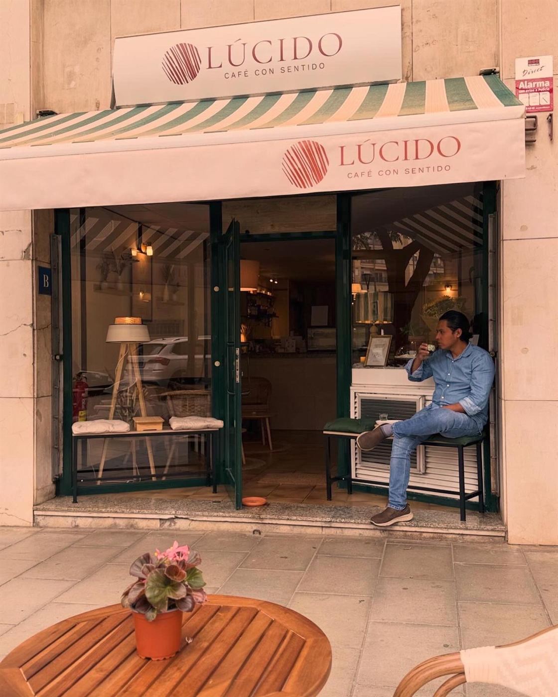 Fachada de Lúcido en Carrer de Camil Fabra: rótulo, toldo a rayas verdes y blancas, puerta abierta y una mesa de la terraza con una planta.
